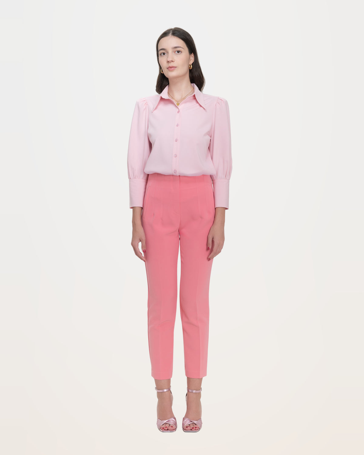 A woman in a pale pink point-collar blouse tucked into coral trousers, standing full length.